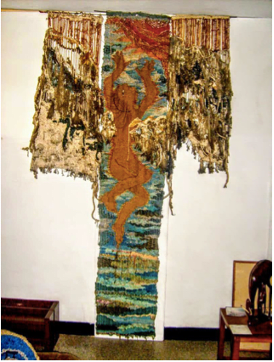 A tapestry hanging against a white wall. I depicts the figure of a woman in a central panel.