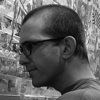 Black and white photograph of a man with glasses. He has a close buzz-cut hair style and looks to be working intently on something off-camera.