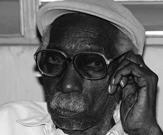 Black and white photograph of a dark-skinned elder man with a thick, white, moustache looking directly at the camera. He wears a flat cap and large-rimmed sunglasses.