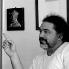 Black and white photograph of a man with a beard and tie-back long hair. He has a raised index-finger as if mid-conversation. 