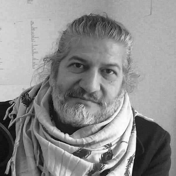 A black and white photograph of a man with a white beard and tied back grey hair. He looks, smiling, at the camera. 