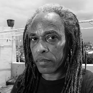 Black and white photograph of a man looking directly at the camera. He has long dreadlocks which are greying at the root.