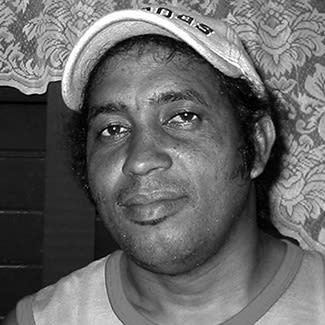 A black and white photograph of a cleanly-shaven dark skinned man. He is smiling slightly and wears a cap. He looks to be early thirties.