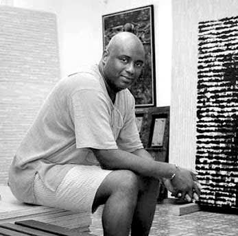 Portrait of a bald dark-skinned man sitting on a bench surrounded by art. He is smiling and wears a T-shirt and shorts.