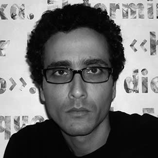 Black and white photograph of a man looking intensely directly at the camera. He is cleanly-shaven and wears squarish glasses. 