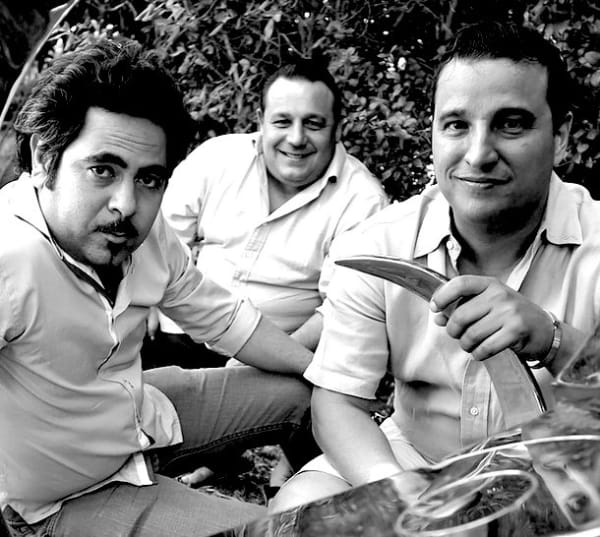 Black and white photograph of three men dressed in white shirts seated on the floor looking directly at the camera. 