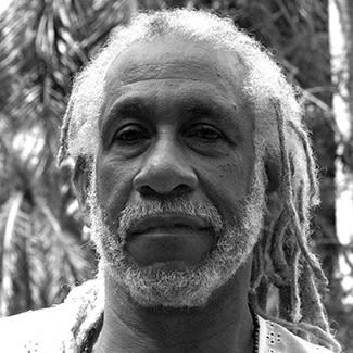 Black and white photograph of a man with a white beard and white dreadlocks looking directly at the camera. his hair is tied back.