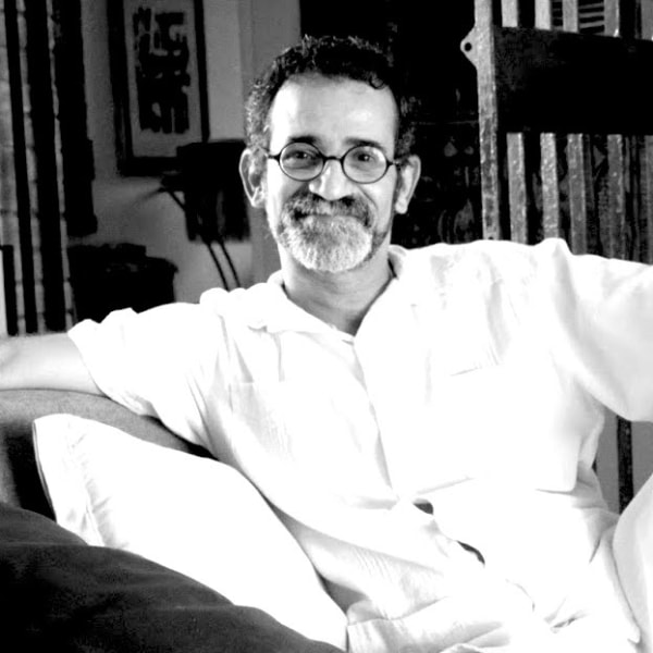 Black and white photograph of a smiling man with a goatee and glasses looking at the camera. He is seated comfortably on a chair and wears thin-rim glasses.