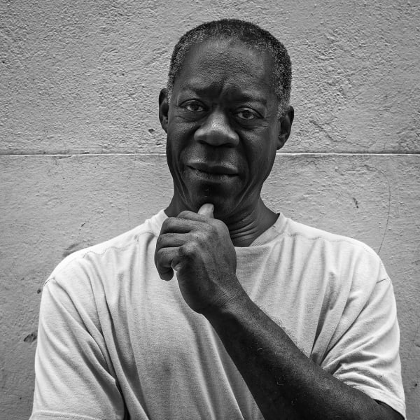 Black and white photograph of a dark-skinned man looking directly at the camera. His hand is raised to his chin in a pose of thoughtfulness. He is cleanly shaven with close-cropped hair. 