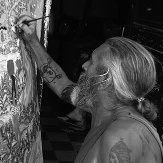 Black and white photograph of a bearded man with long, tied-back hair painting on a canvas. He is facing away from the camera.