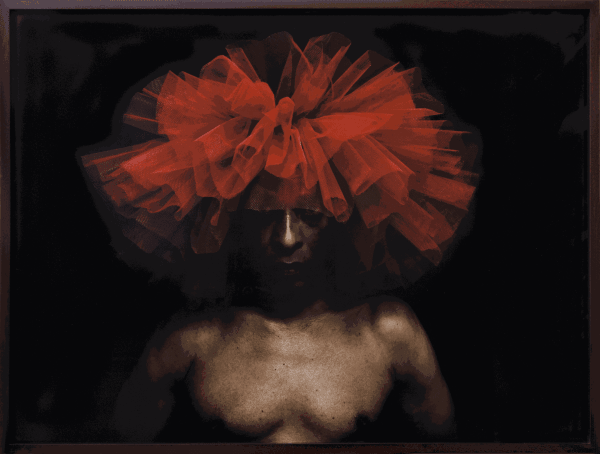 The portrait of a dark-skinned man wearing a bright red tutu as a headdress. He is photographed against a black background.